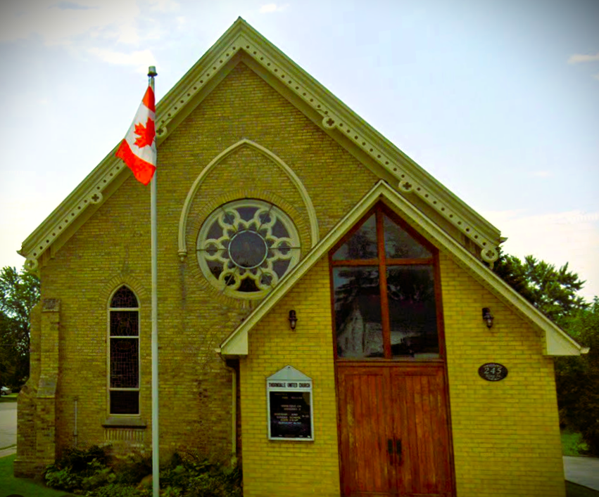 a yellow brick church with wood doors in the foreground a flagpole with a canadian flag and in the background blue sky and trees