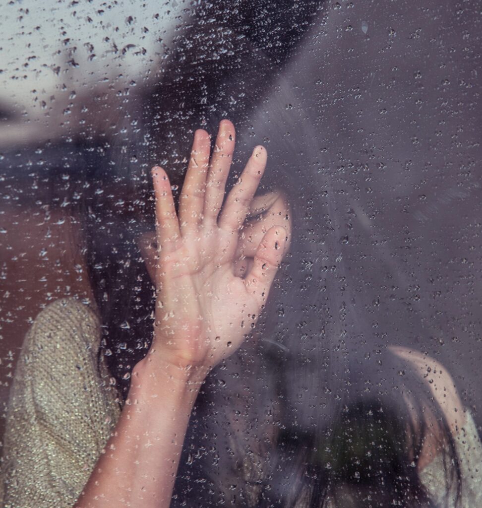 a glass window covered in rain in the background a woman is holding her hand on the glass of the window
