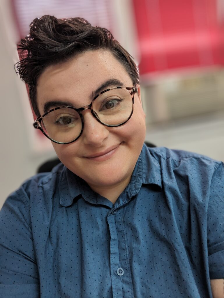 a person with short dark hair wearing glasses and smiling they are wearing a blue button down shirt in the back ground is a pink and red display