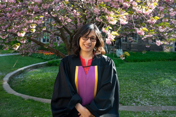 a woman with shoulder length brown hair wearing glasses and smiling, she is wear a black robe with a pink and yellow dress underneath, in the background is a blooming magnolia tree