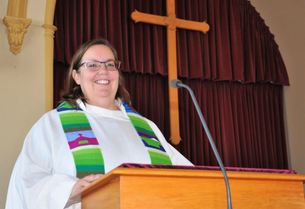 a woman with long brown hair, wearing glasses and smiling at the camera wearing white vestments and a green stole, she is standing at a wood lectern in the back ground are burgundy drapes and a wood cross